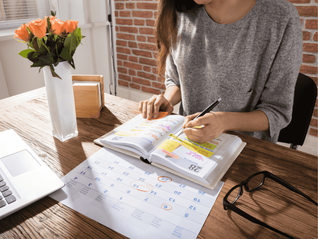 A woman is sitting at her desk, and writing in her calendar book. There are different items surrounding her, like her glasses, a monthly overview, and a block with little notes. - long-term blogging goals