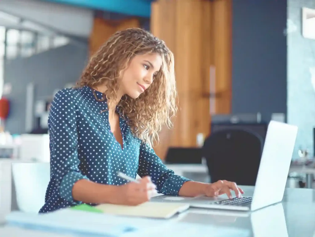 A young woman with curly hair works diligently at her laptop while taking notes, showcasing focused and productive work habits in a modern office setting. - habits to boost productivity