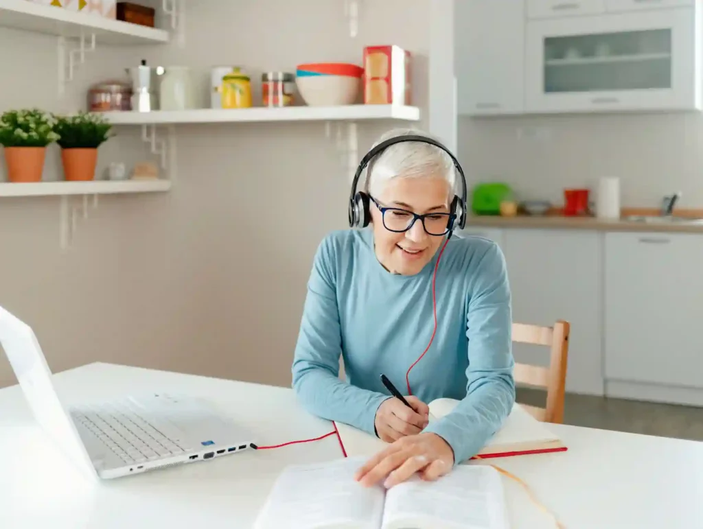 A smiling woman with short gray hair and glasses sits at a kitchen table, wearing headphones and writing in a notebook while working on a laptop, demonstrating focused productivity in a bright, organized home environment. - habits to boost productivity