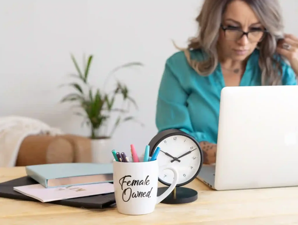 A woman with glasses and blonde hair works intently on a laptop, surrounded by productivity tools including a "Female Owned" mug, colorful pens, and a desk clock - time tracking tools.