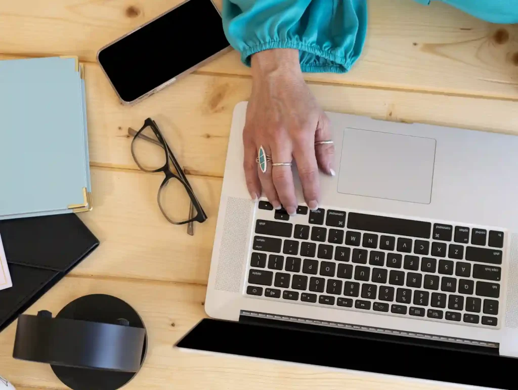 A close-up of a woman's hand typing on a laptop keyboard, with a smartphone, glasses, and a desk clock visible on the wooden desk - time tracking tools.