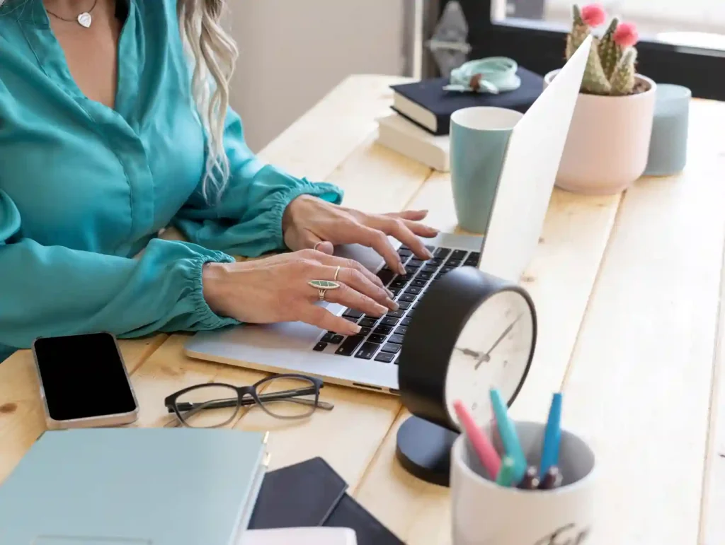 A woman in a teal blouse types on a laptop, with a desk organized with a clock, colorful pens, glasses, and a cactus plant nearby - time tracking tools.