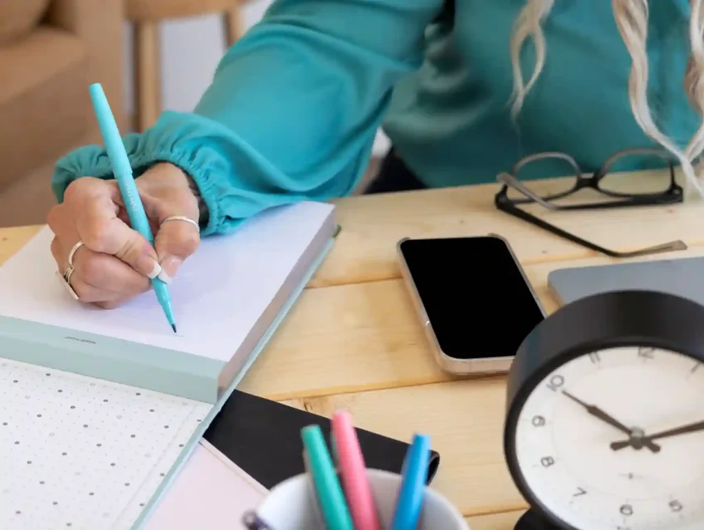 A woman in a teal shirt writes in a notebook with a blue pen, surrounded by productivity essentials including a smartphone, glasses, and a desk clock - time tracking tools.