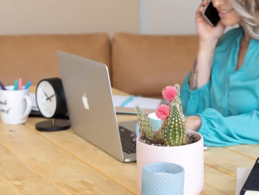 A woman in teal talks on the phone near her laptop, with a workspace featuring a clock, colorful pens, and a blooming cactus plant - time tracking tools.