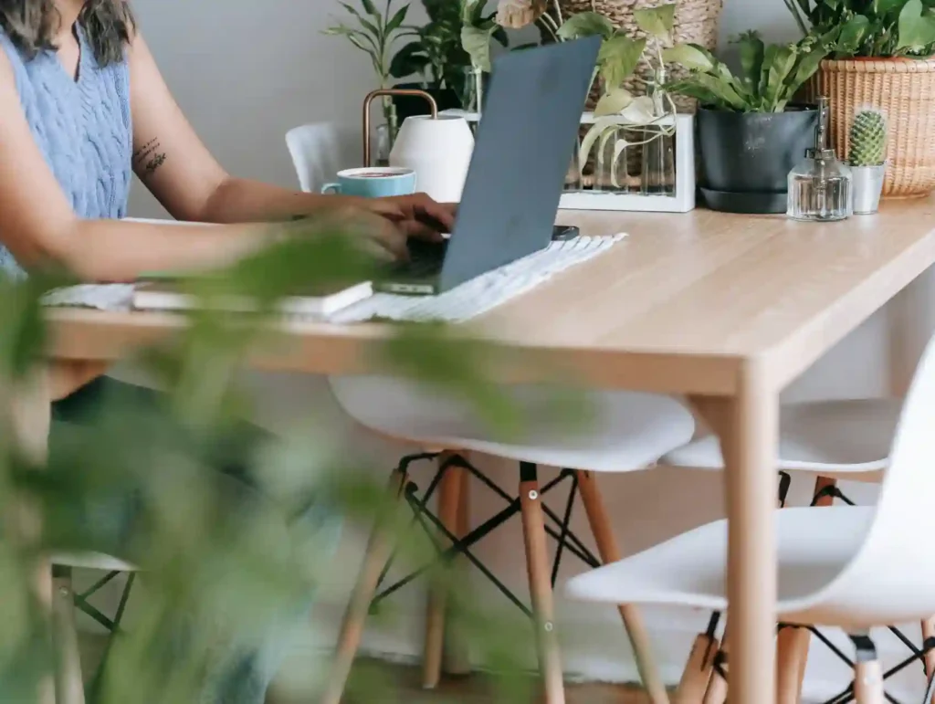 A woman types on a laptop in a plant-filled workspace, surrounded by potted greenery and personal decor, embodying a focused and nature-inspired work environment. - monotasking habit