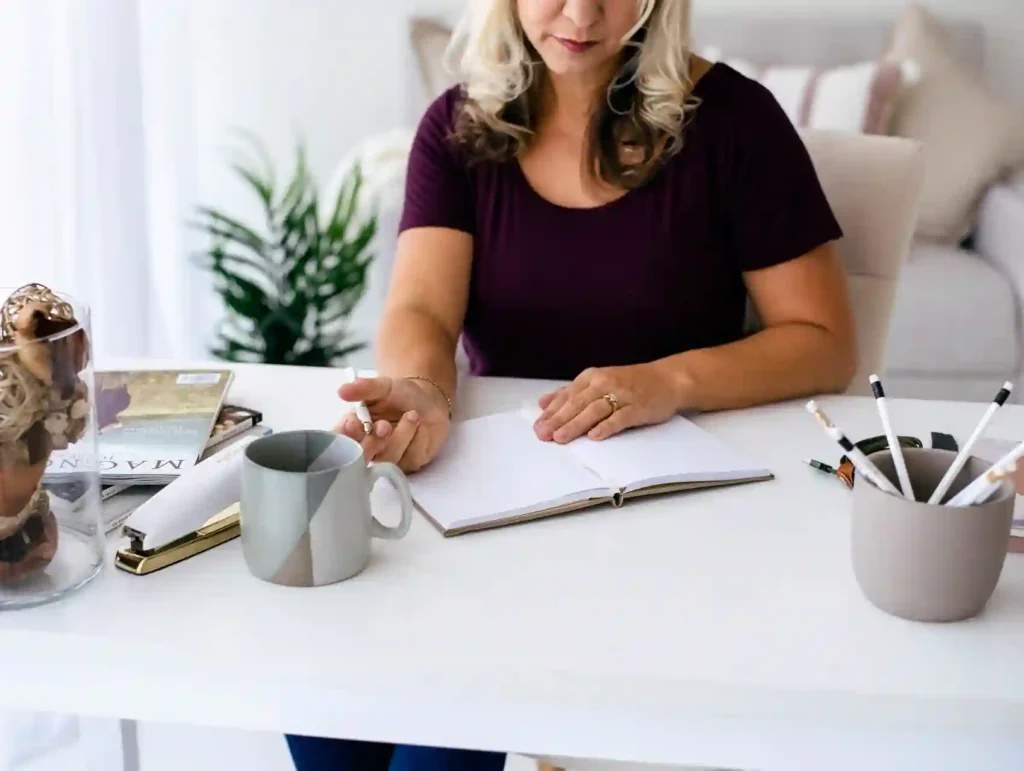 A close-up view of a woman's hands writing in an open notebook on a white desk, with a coffee mug and pens visible nearby. - habit of planning ahead
