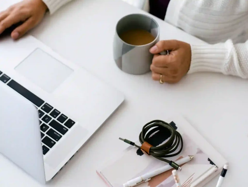 A woman's hands hold a gray mug of coffee next to a laptop, with a coiled charging cable and pens on a white desk, showcasing a tidy workspace. - habit of planning ahead