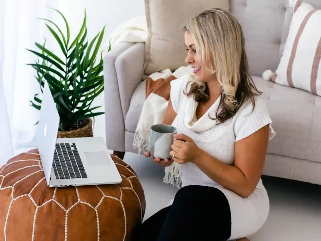 A smiling blonde woman in a white top sits on the floor, holding a coffee mug while working on a laptop perched on a brown pouf, with a potted plant nearby. - habit of planning ahead