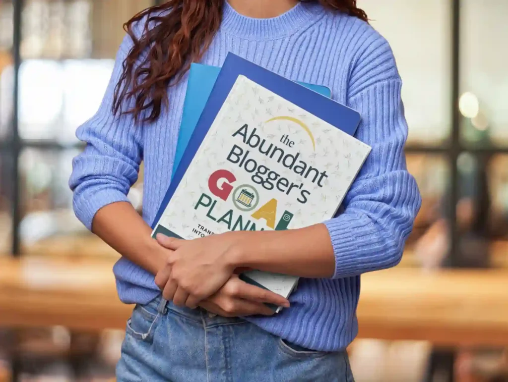 A woman wearing a light blue ribbed sweater and jeans holds "The Abundant Blogger's GOAL PLANNER" book along with other blue folders against their chest in an indoor setting with a blurred background.