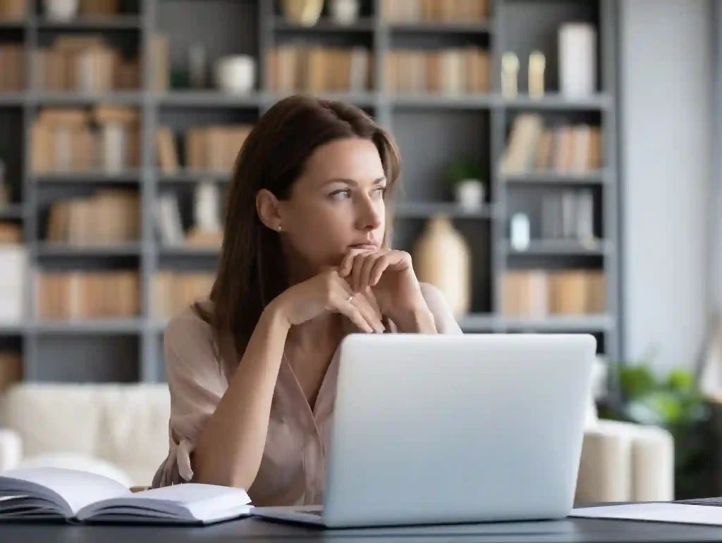 A thoughtful woman in a beige blouse sits at a desk with a laptop and open notebook, with a modern bookshelf filled with tan-colored books visible in the background - why store blog post ideas