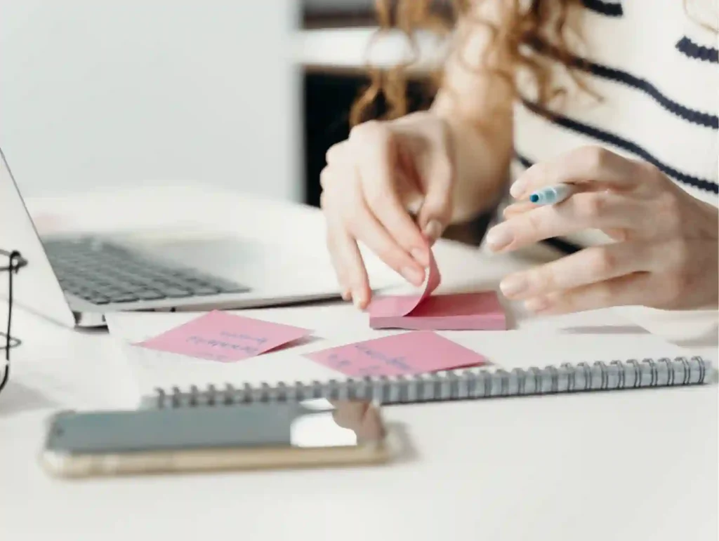 A close-up view of hands arranging pink sticky notes on a spiral notebook next to a laptop, showing the process of organizing ideas - why store blog post ideas