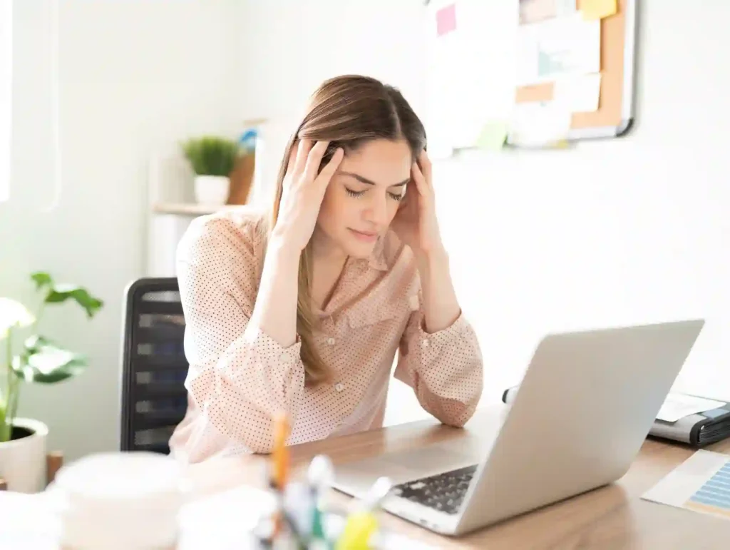 A woman in a peach-colored polka dot blouse sits at a desk with a laptop, holding their head in apparent frustration while working - why store blog post ideas