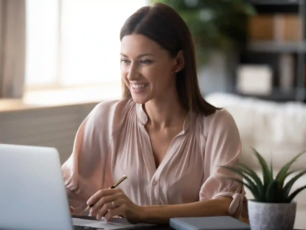 A smiling woman in a blush-colored blouse sits at a desk with a laptop and small potted plant, appearing engaged and productive in her work environment - why store blog post ideas