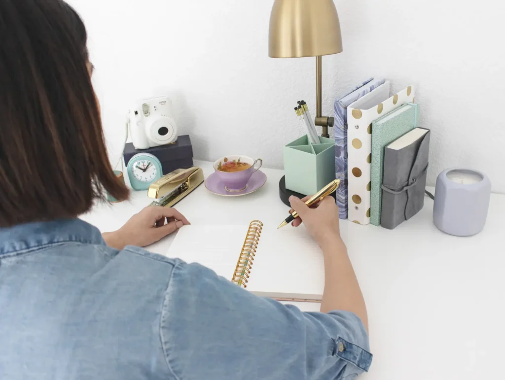 A woman wearing a denim shirt sits at a white desk with a gold lamp, writing in a spiral notebook surrounded by organized stationery and a purple teacup - journaling for productivity