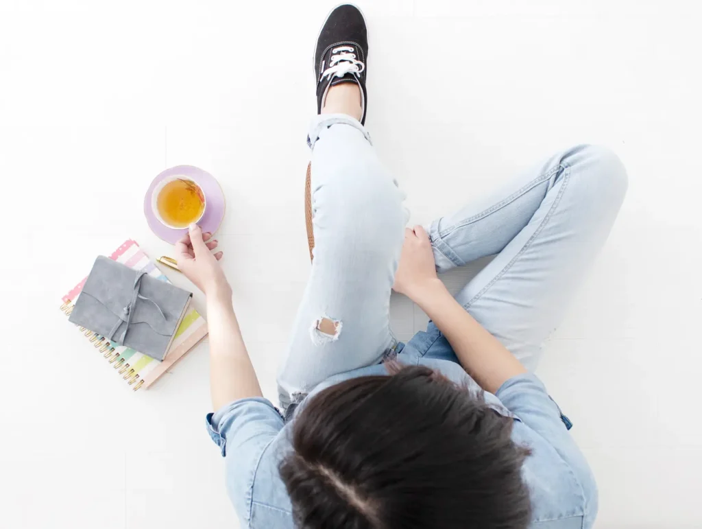 An overhead flatlay of a woman wearing distressed light denim and black sneakers, sitting cross-legged with a pink notebook and lavender teacup on a white background - journaling for productivity