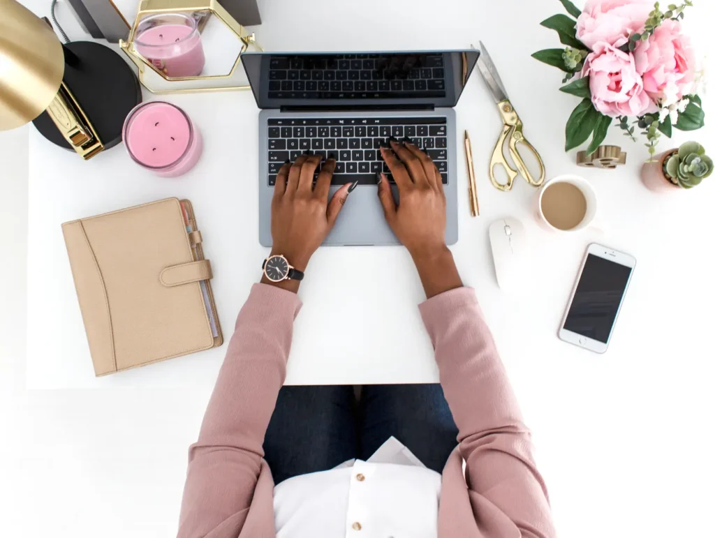Hands type on a MacBook keyboard while wearing a pink blazer, with a beige planner, pink candles, gold scissors, and fresh flowers arranged on a white desk - time management vs task management
