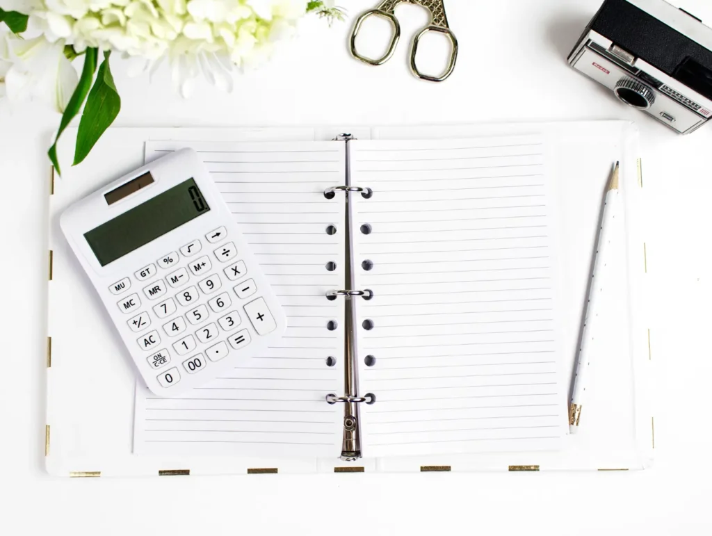 An open ring-bound planner with lined pages lies flat beside a white calculator, decorative scissors, and white flowers - brain-dump