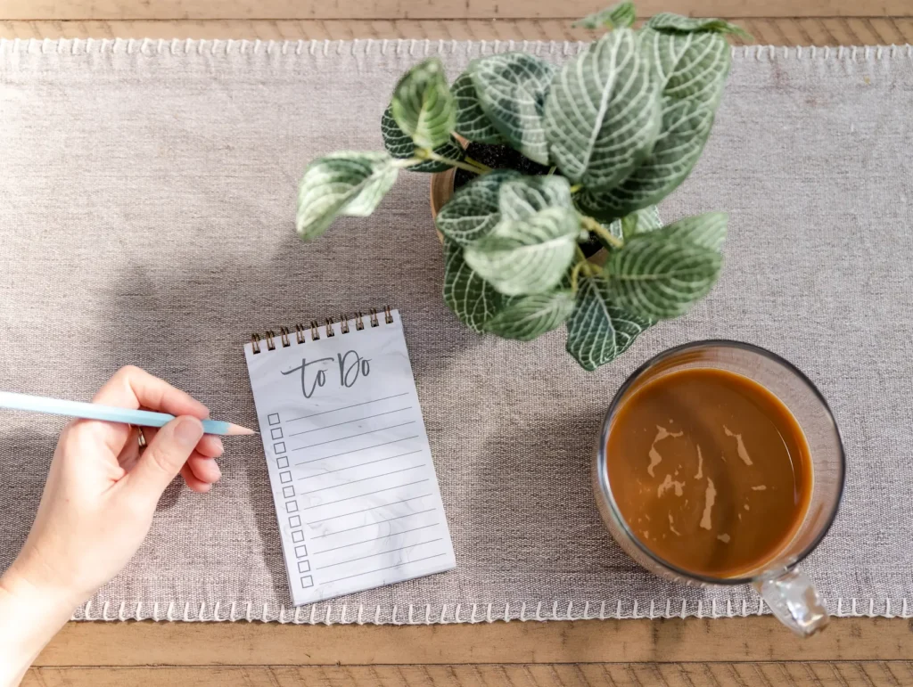 A hand holds a blue pen over a spiral "to do" notepad, with a houseplant and coffee cup arranged on a textured placemat - pockets of time