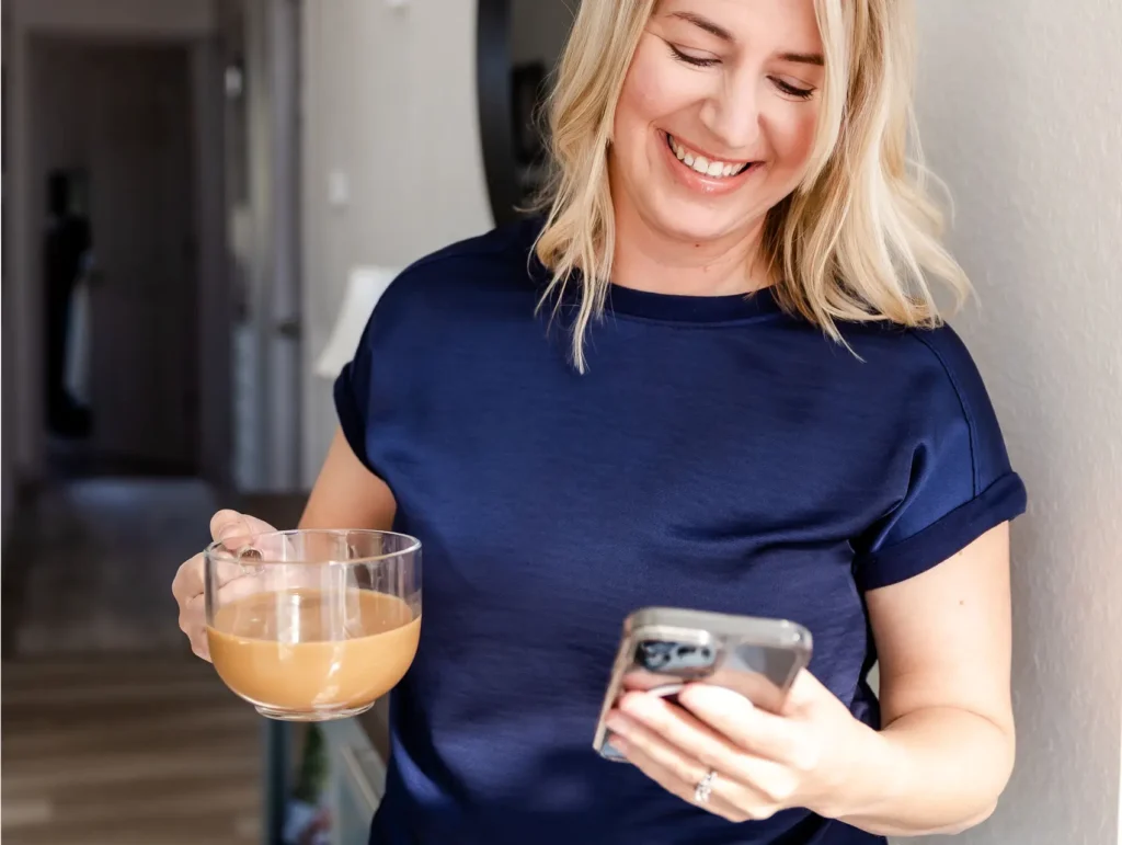 A person in a navy blue t-shirt smiles warmly while holding a glass of creamy coffee in one hand and checking their phone in the other - pockets of time