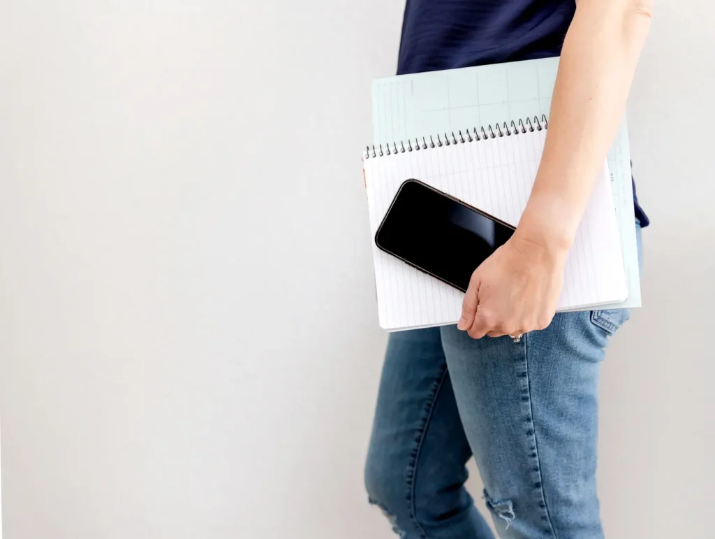 A person wearing a navy blue shirt and distressed jeans holds a spiral notebook and smartphone against a minimalist white wall - pockets of time