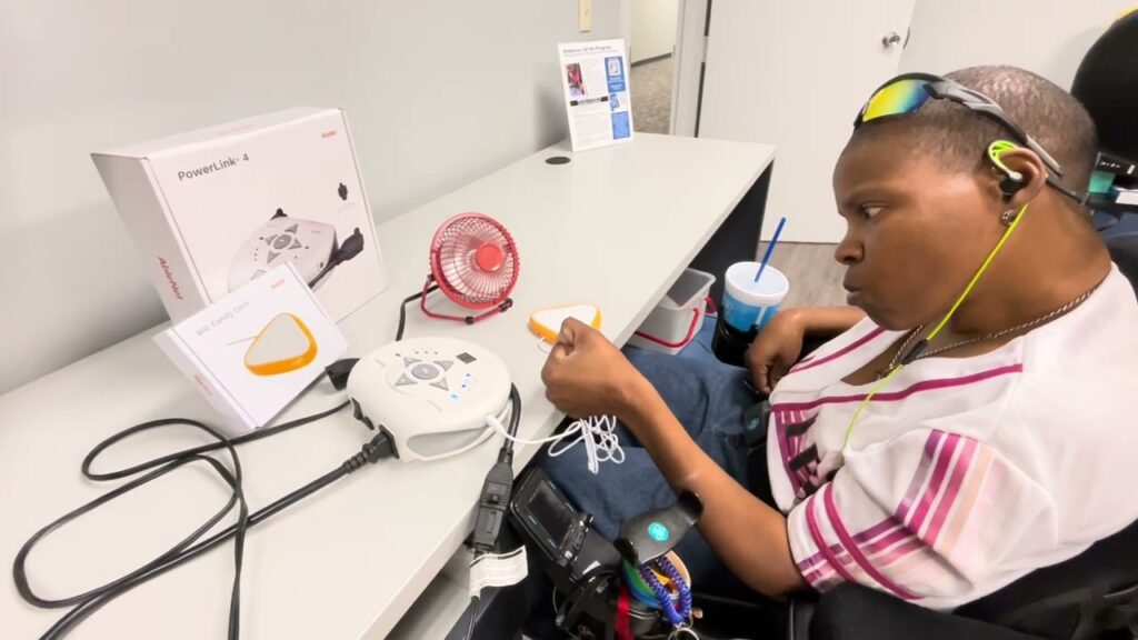 Nakisha uses an adaptive device with push buttons to control a small red fan on a table, with a drink and instruction booklet nearby.