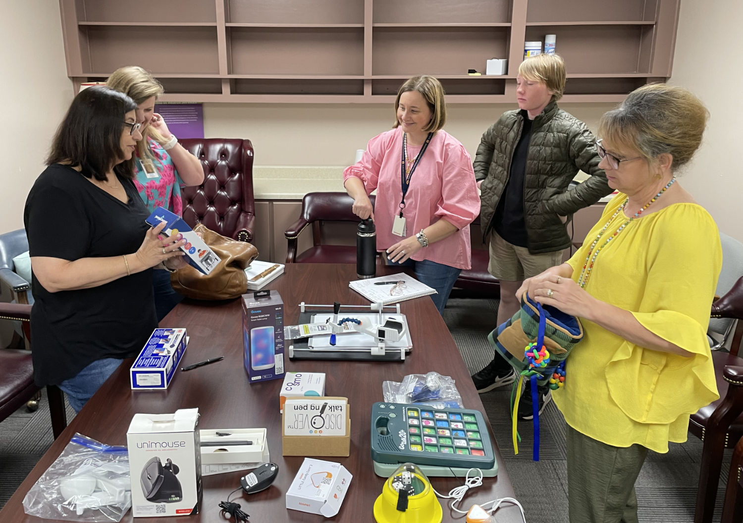 Five people stand around a table covered with various electronic devices and assistive technology items in an office setting.