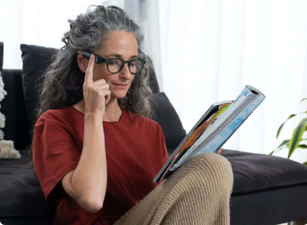 A woman with gray hair and glasses sits on the floor reading a magazine and touching the Orcam device on her glasses with one finger.