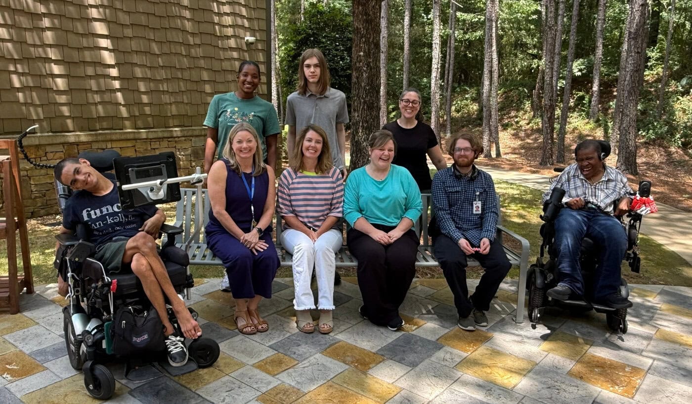 APTAT team: a group of 9 adults, including two in wheelchairs, pose outdoors on a paved area in front of trees and a building.