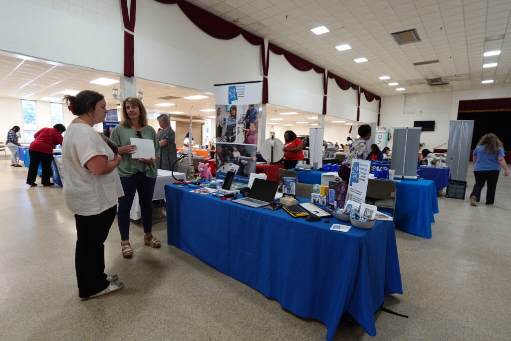 A woman speaks with an exhibitor at a table displaying various assistive tech