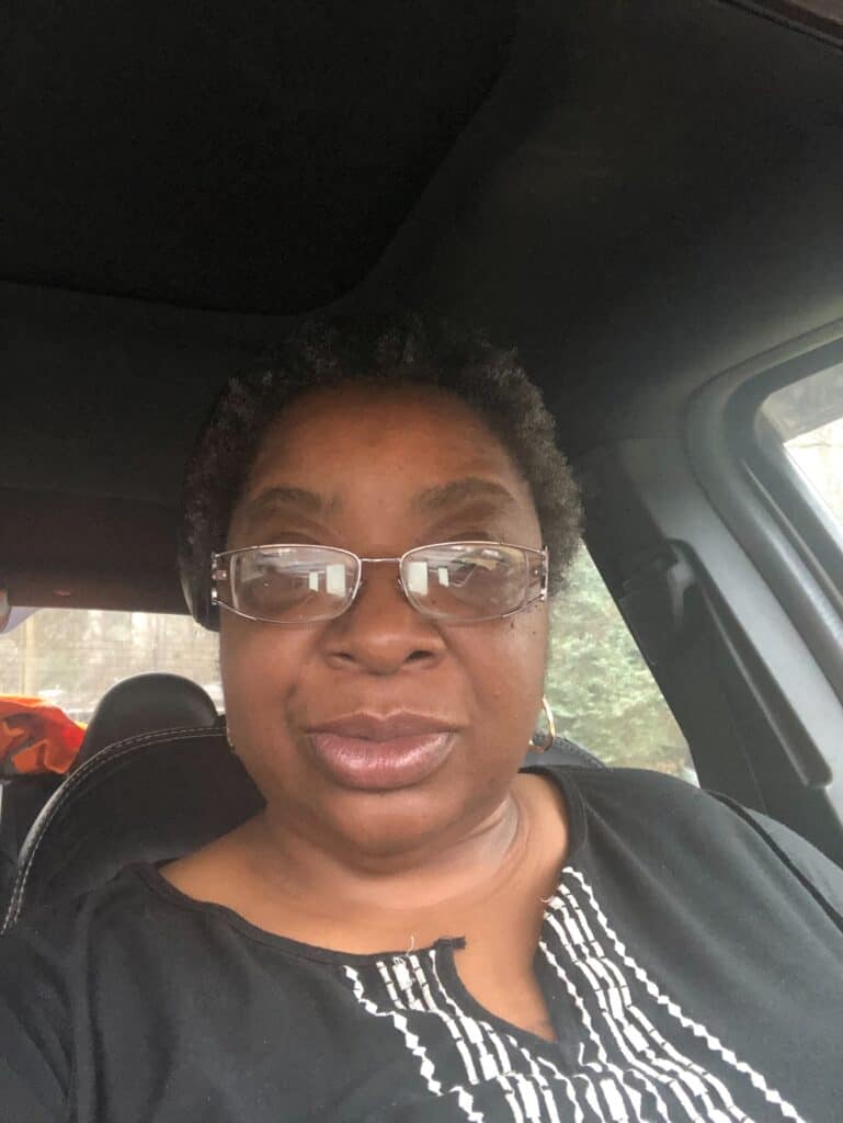 Sharon Henderson is wearing glasses and a black and white top, sitting inside a car.