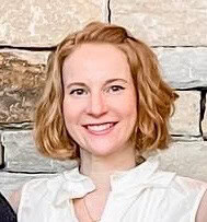 Virginia Rediker has short, wavy, light brown hair, is smiling, wearing a white top and standing in front of a stone wall.