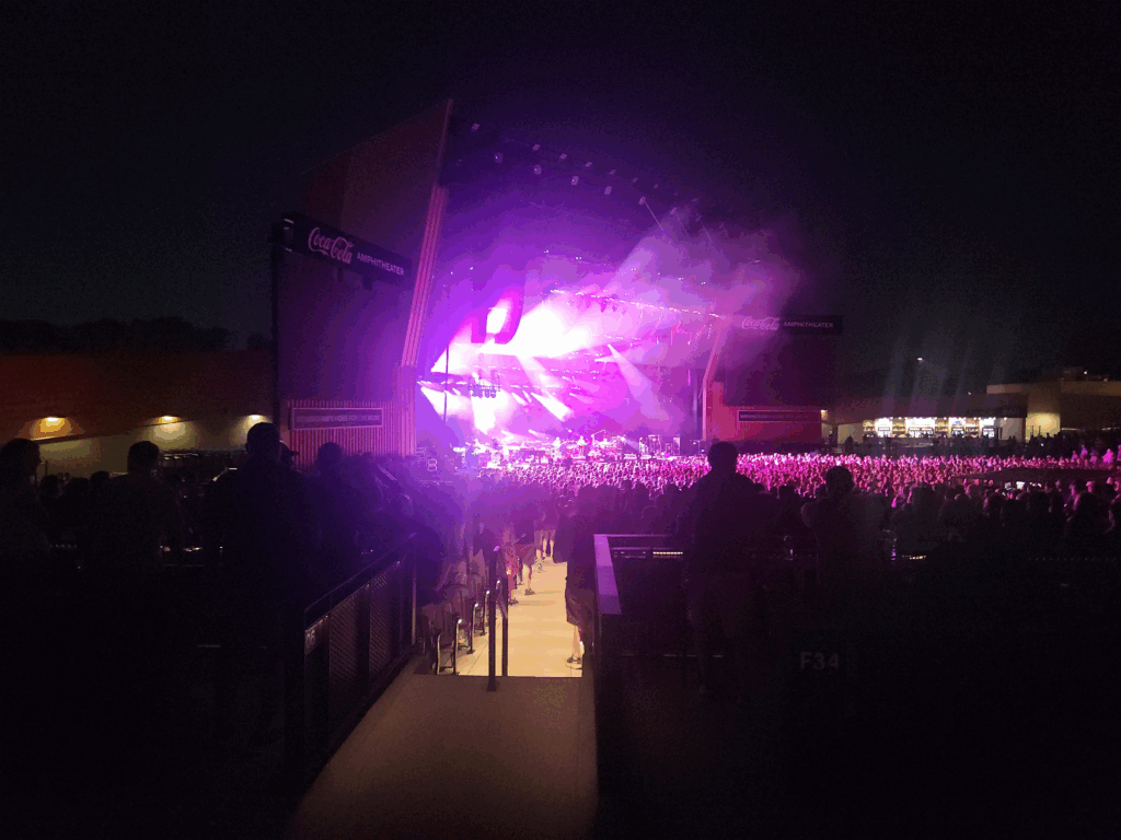 Vast outdoor concert at night with a large crowd facing a brightly lit stage with purple and white lights, and a giant "CocaCola Amphitheater" sign on the side.