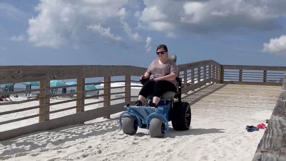 Ashley driving a powered beach wheelchair from a wooden boardwalk ramp onto the sand at Gulf State Park