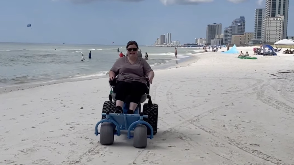 Ashley driving a powered beach wheelchair along the shoreline at Gulf State Park with ocean and beachgoers in the background