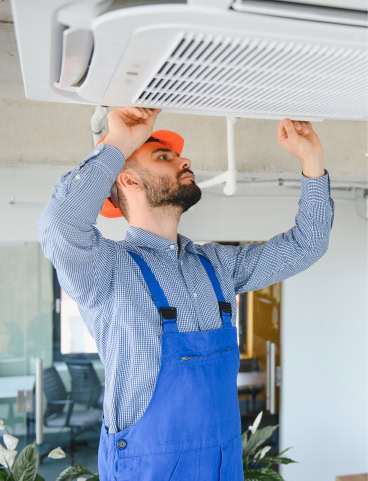 Man in orange hard hat and blue overalls installing a ceiling air conditioner.
