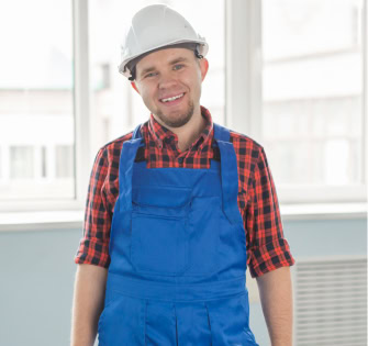 Smiling man in white hard hat, red plaid shirt, and blue overalls.