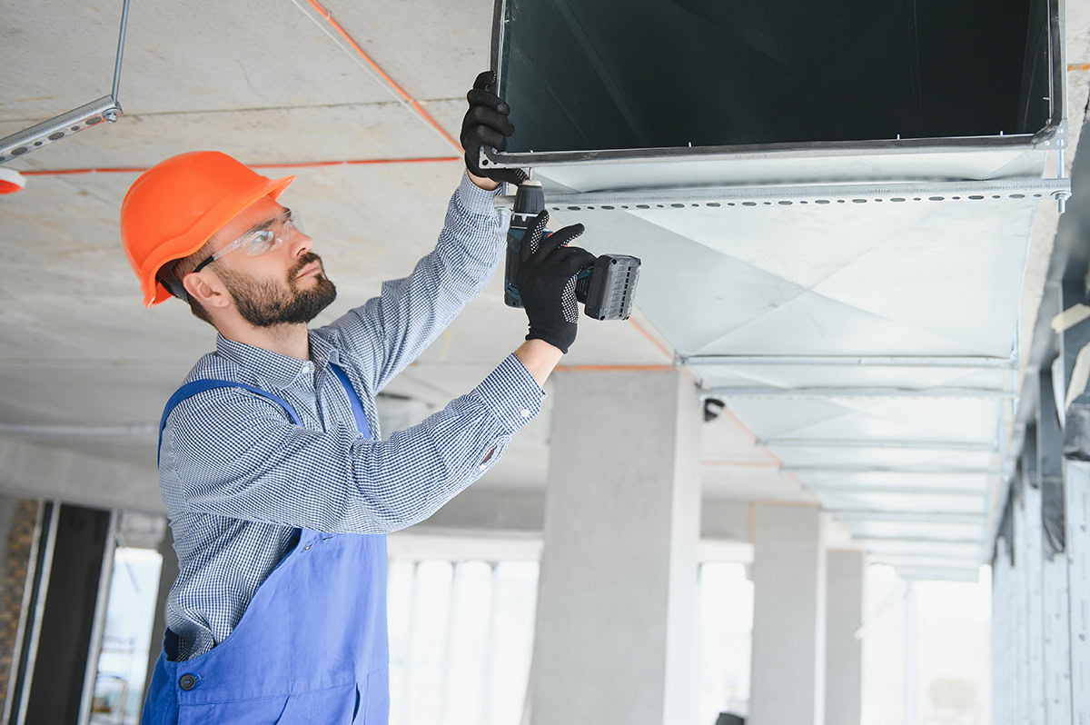 Man in hard hat installing ventilation ductwork with a drill