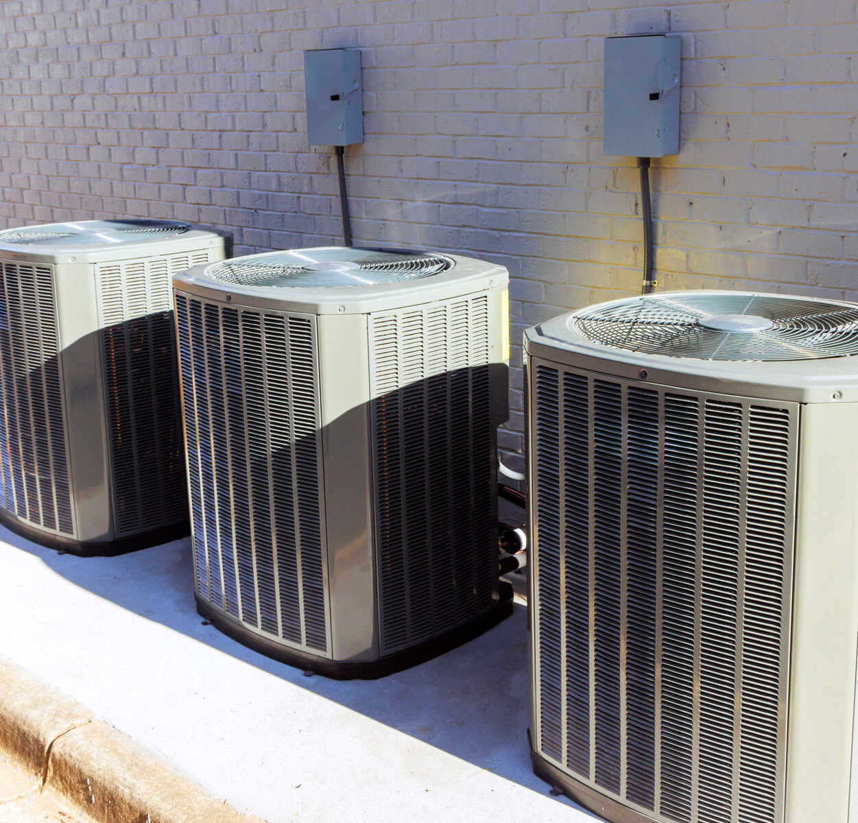 Three outdoor air conditioning units next to a brick wall.
