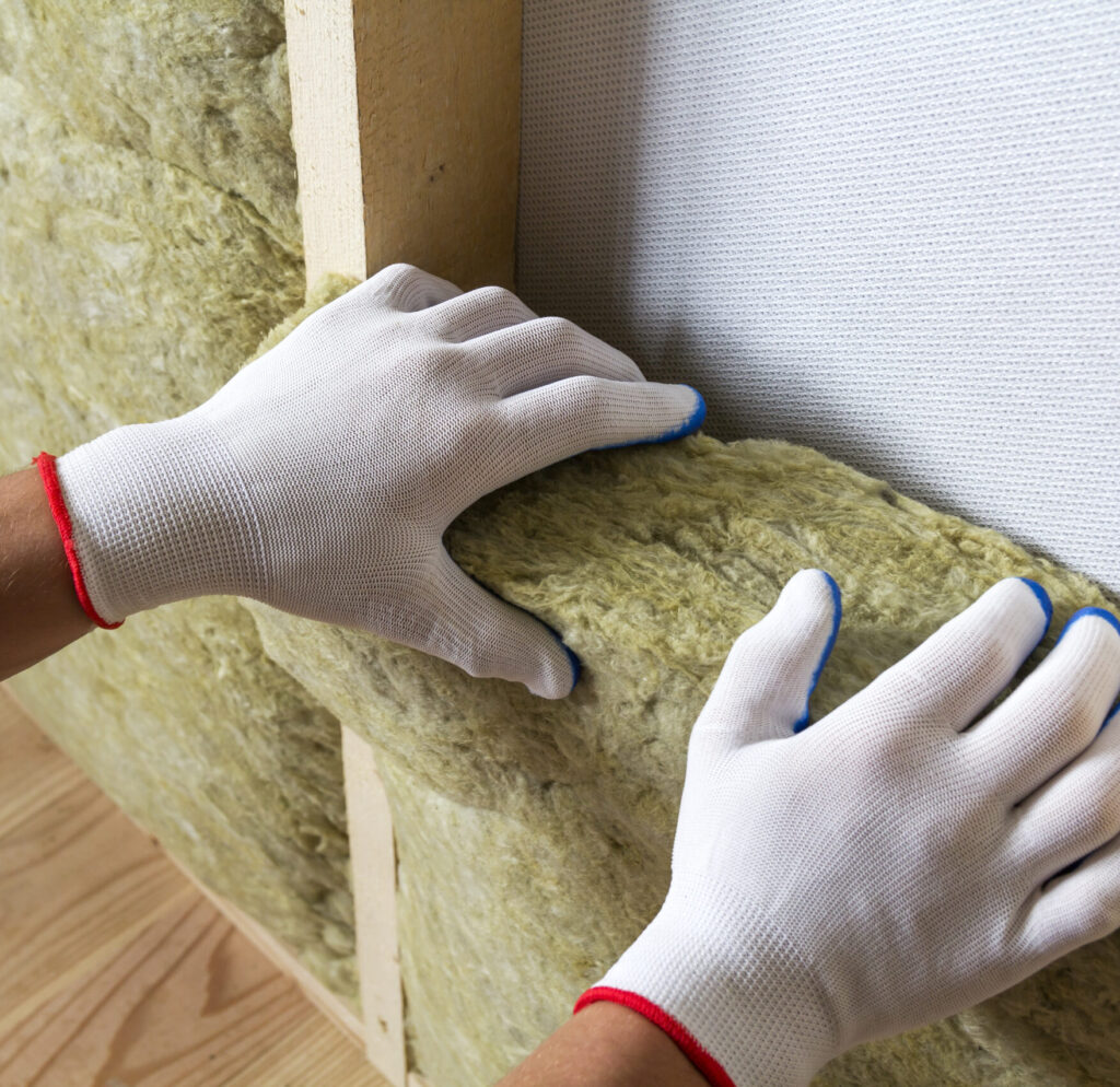 Hands in white gloves installing insulation between wooden studs and a wall