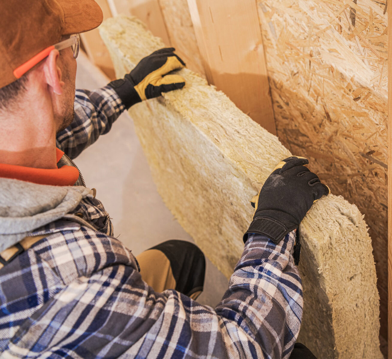 Man in plaid shirt and gloves installing insulation in a wall.