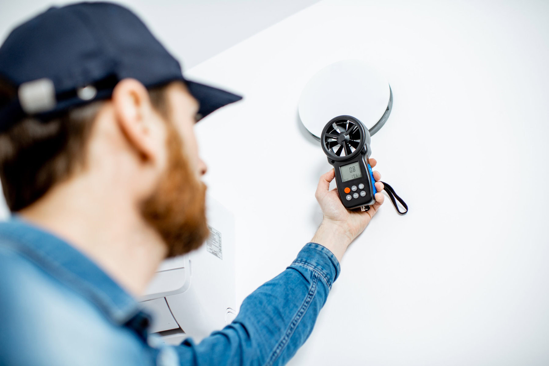 Man in a cap holding an anemometer near a white ceiling vent