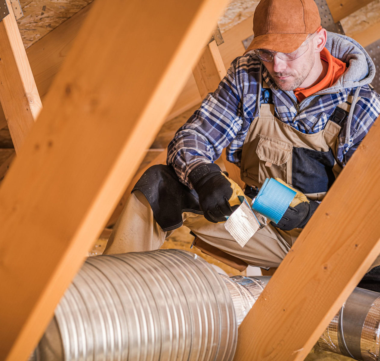 Man in attic installing ductwork, wearing cap, glasses, and work clothes.