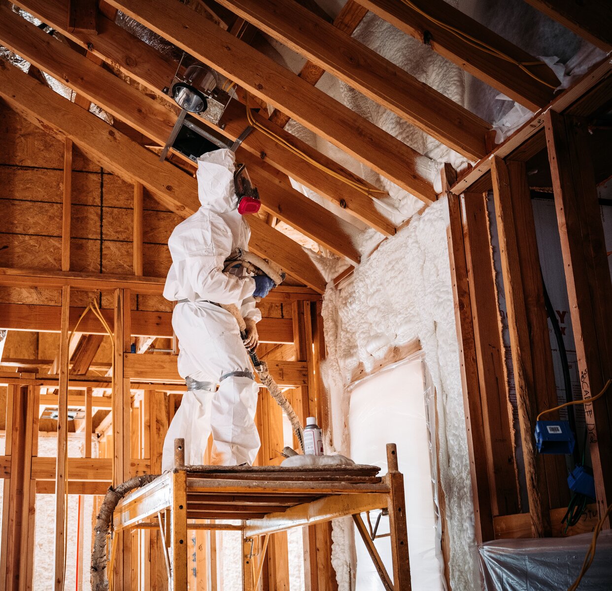 Worker in hazmat suit applying spray foam insulation to a wooden attic.