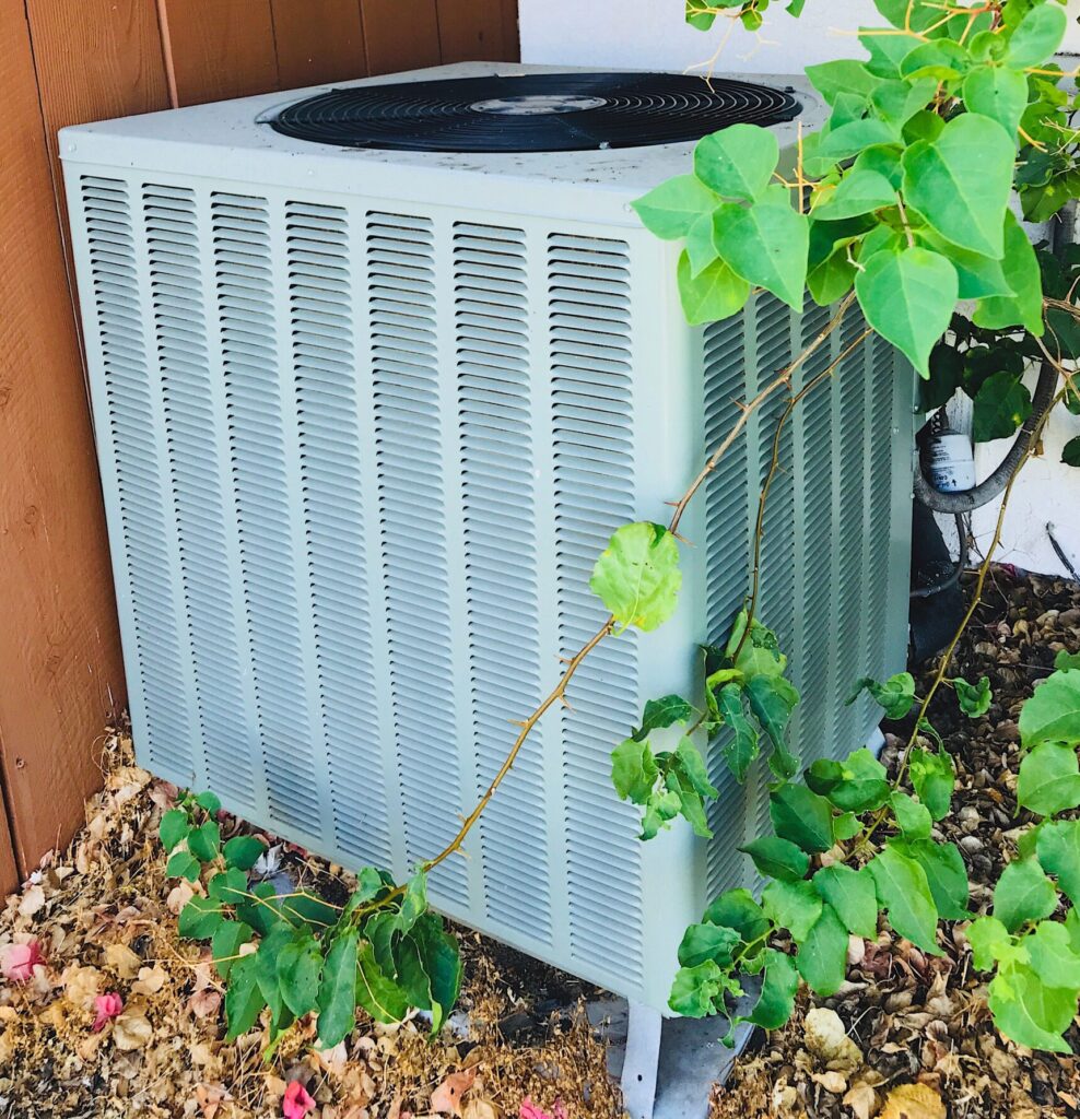 Outdoor air conditioning unit next to a wooden fence and green foliage.