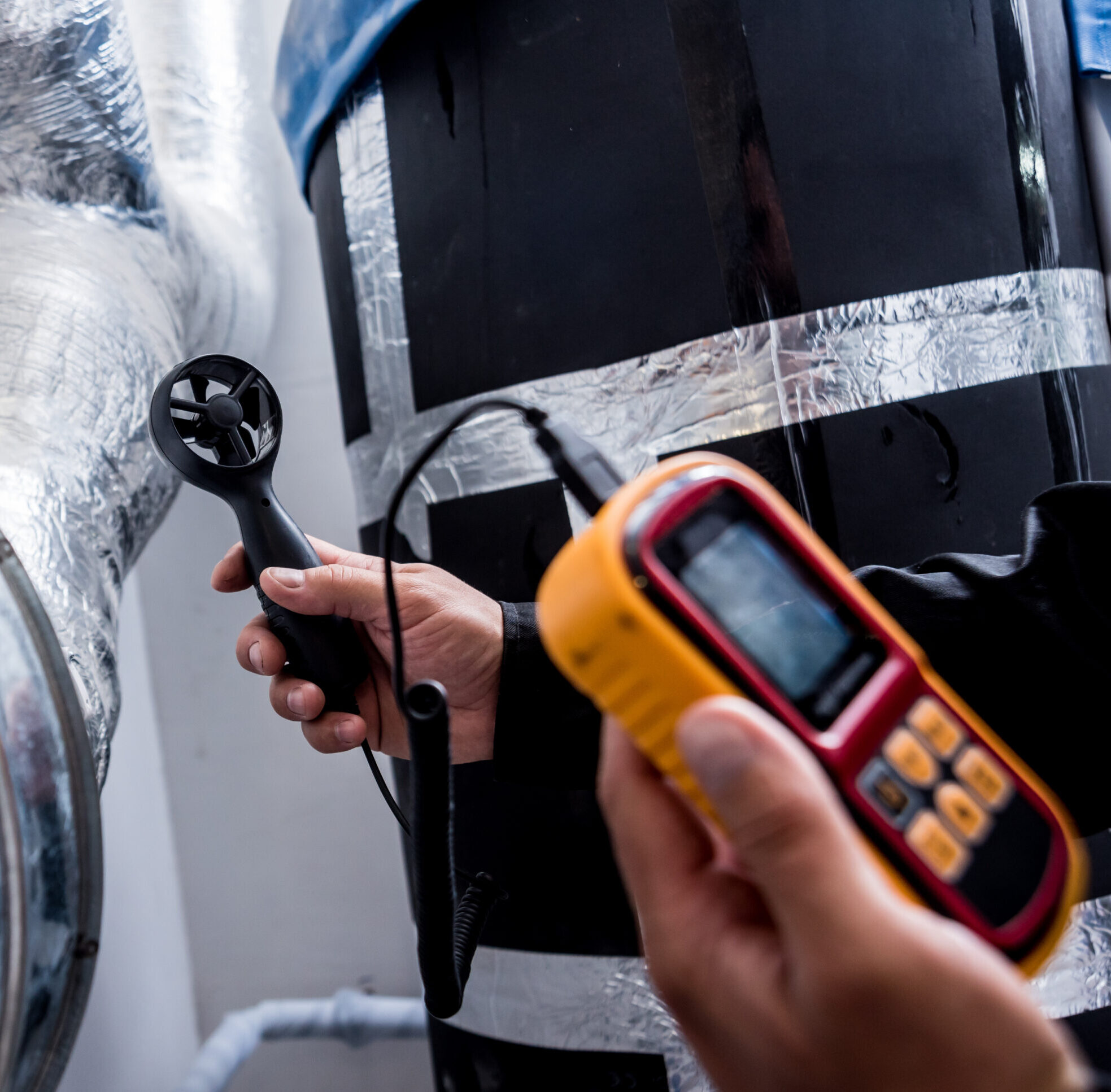 Hands holding an anemometer and a yellow digital meter near insulated pipes.