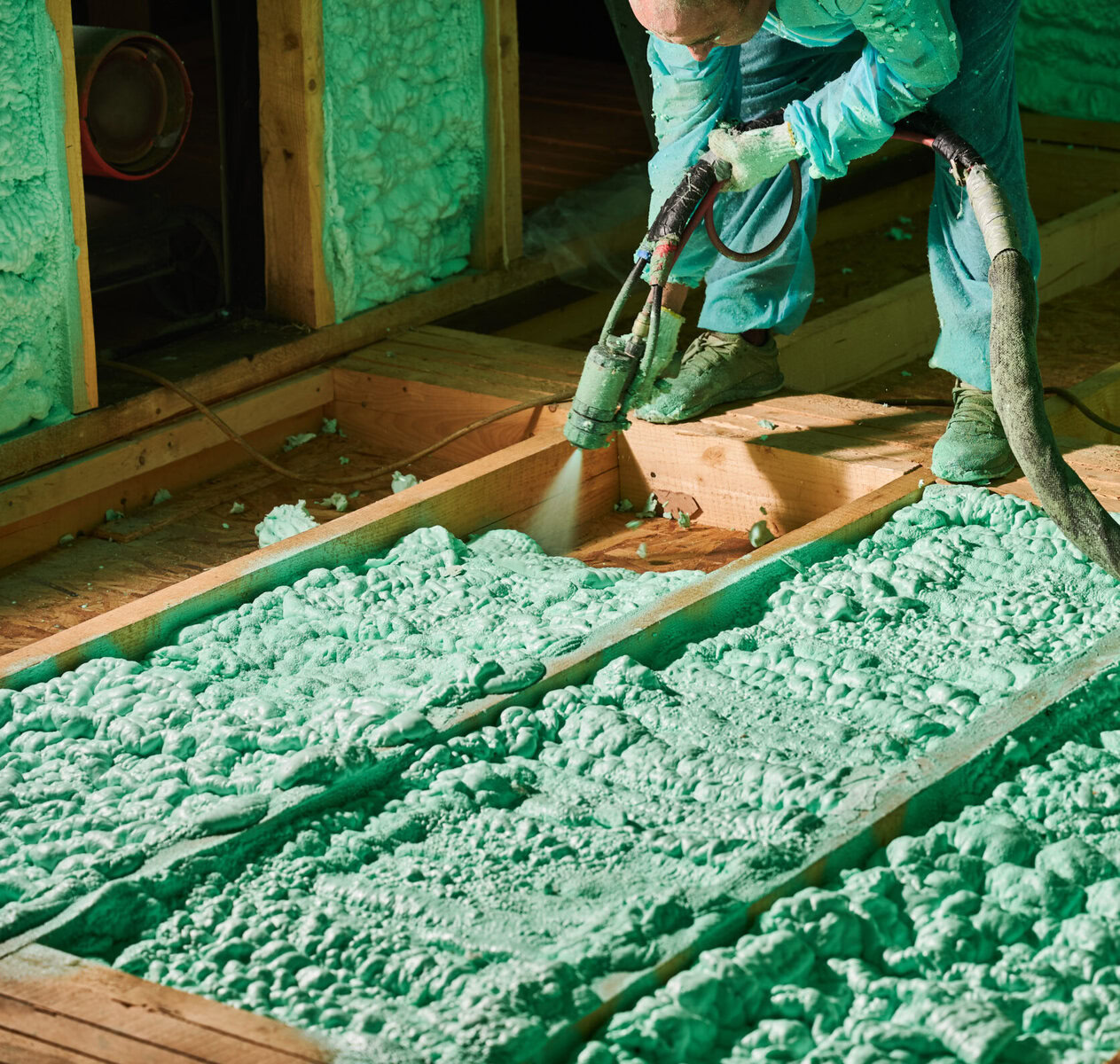 Worker applying green spray foam insulation between wooden joists
