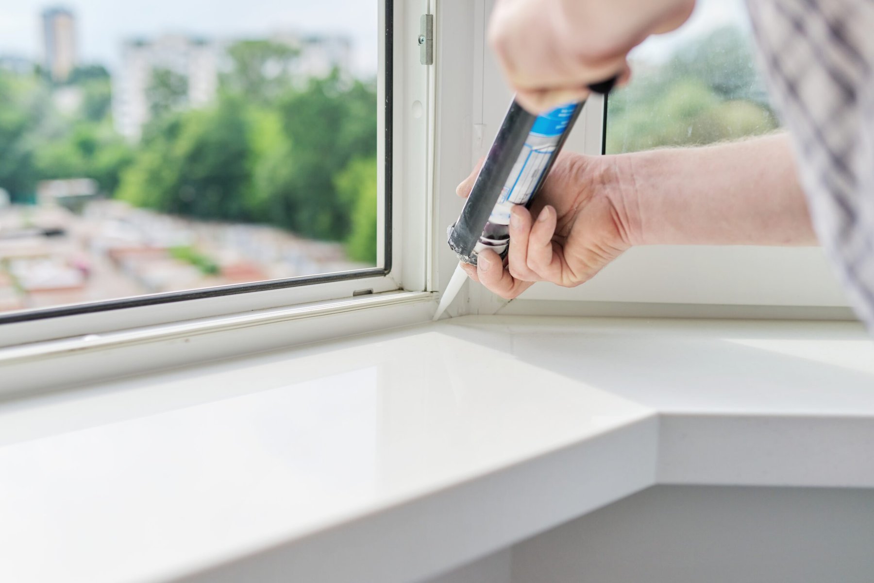 Person applying sealant to a white window frame with a caulking gun.
