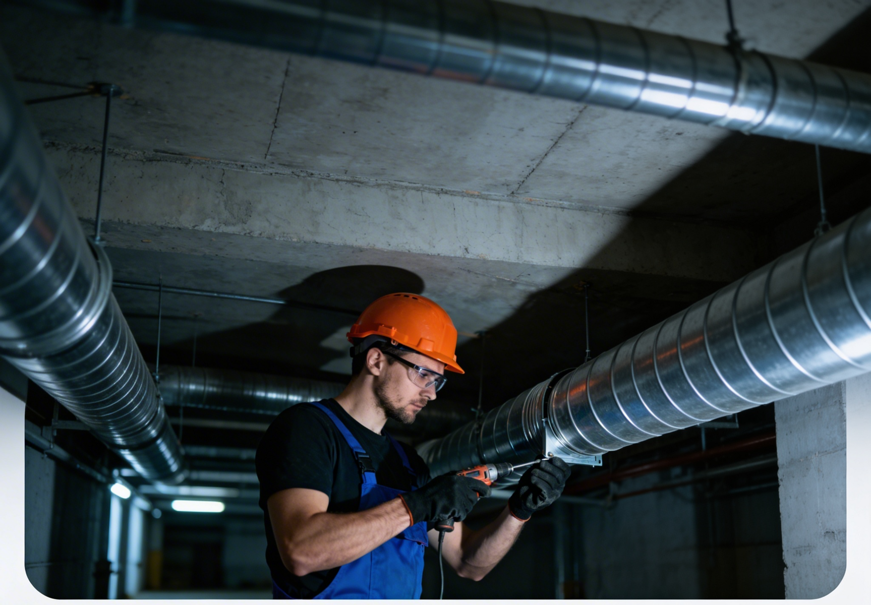 Man in hard hat and glasses working on ventilation pipes with a drill.