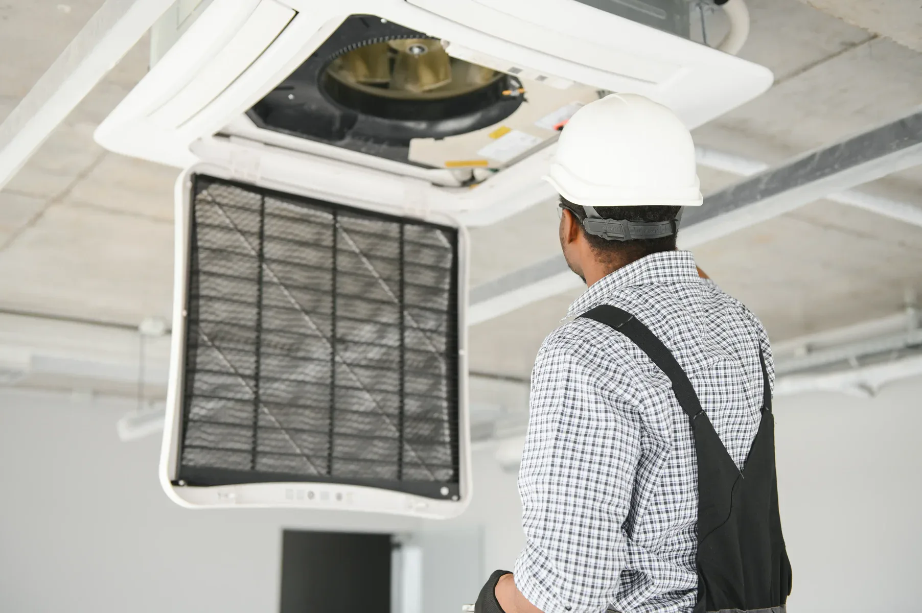 Man in hard hat inspecting a ceiling-mounted air conditioning unit.