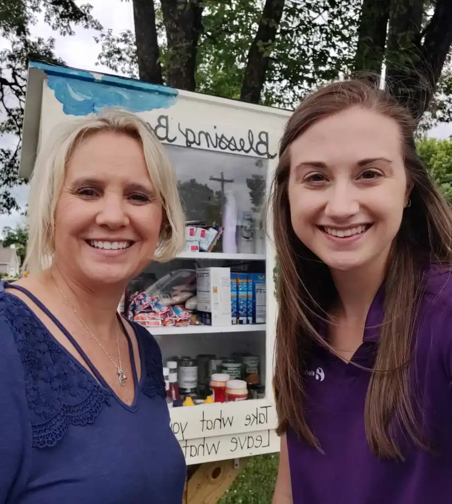 Two women smiling near a Blessing Box with supplies in a park setting.